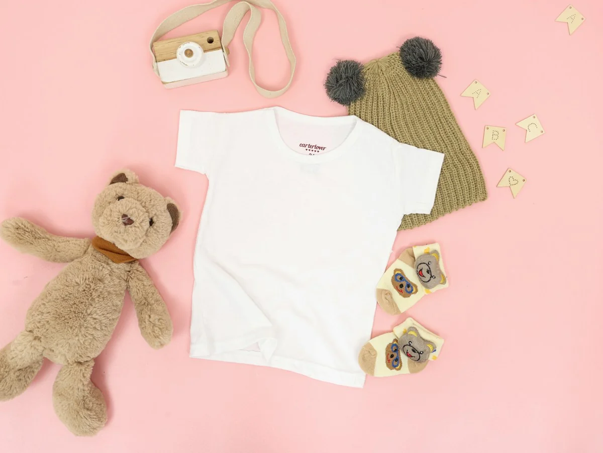Baby clothing, a knitted hat, and a teddy bear arranged on a soft pink background